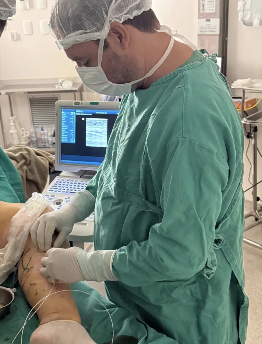 Dr. Alexandre, de touca, máscara e avental cirúrgico verde, faz um procedimento na perna de um paciente guiado pela imagem do ultrassom, visível no monitor ao fundo.